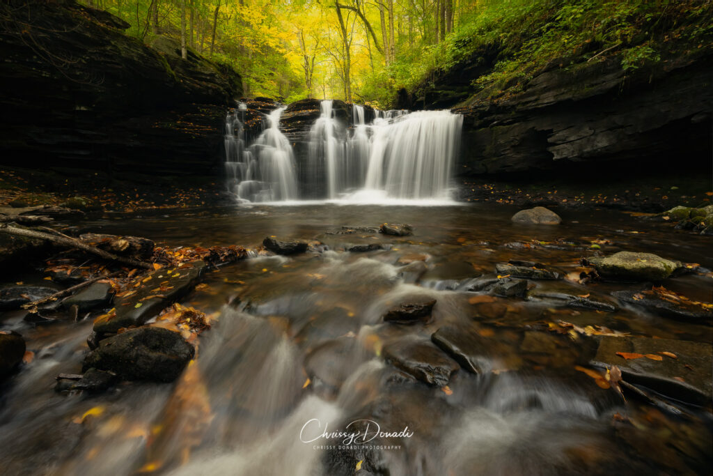 Autumn waterfall in Appalachia in Pennsylvania with yellow and green trees above and orange leaves scattered on the rocks of the creek